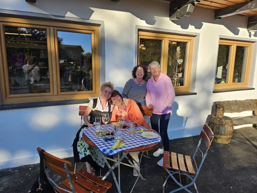 Vier Frauen stehen um einen Tisch im Freien, lachen und genießen die Zeit zusammen.